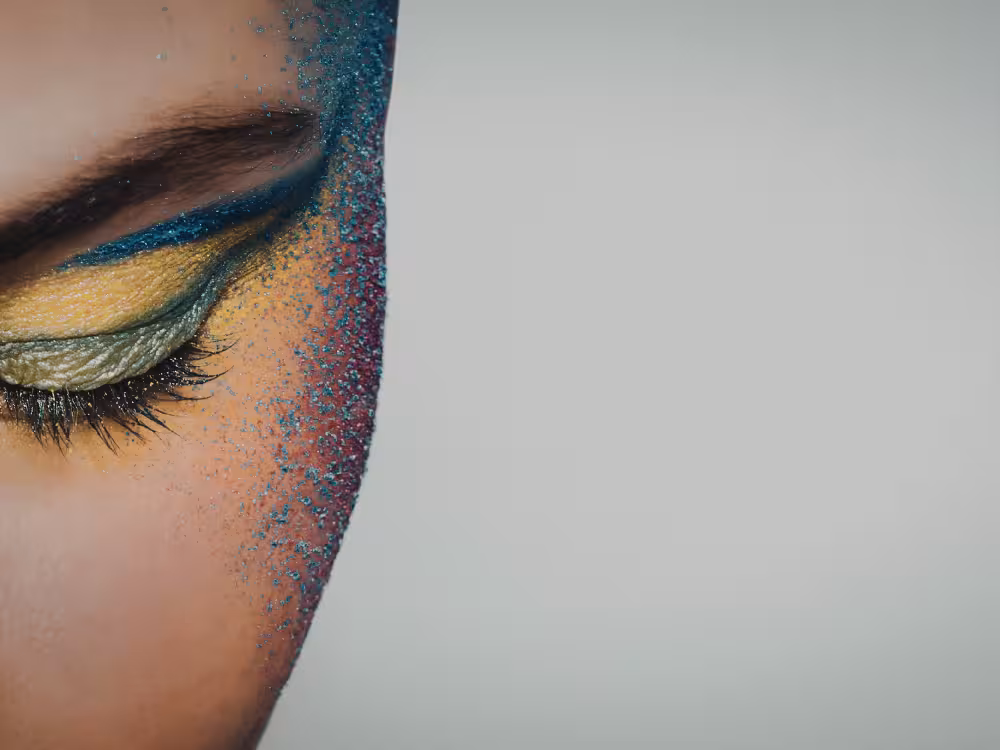 Metade do rosto de uma pessoa com maquiagem artística intensa: sombra amarela dourada na pálpebra com delineador azul-petróleo e glitter azul espalhado por toda a lateral do rosto, sobre uma base em tom vinho. Olho fechado com cílios naturais. Fundo cinza claro com amplo espaço negativo à direita.