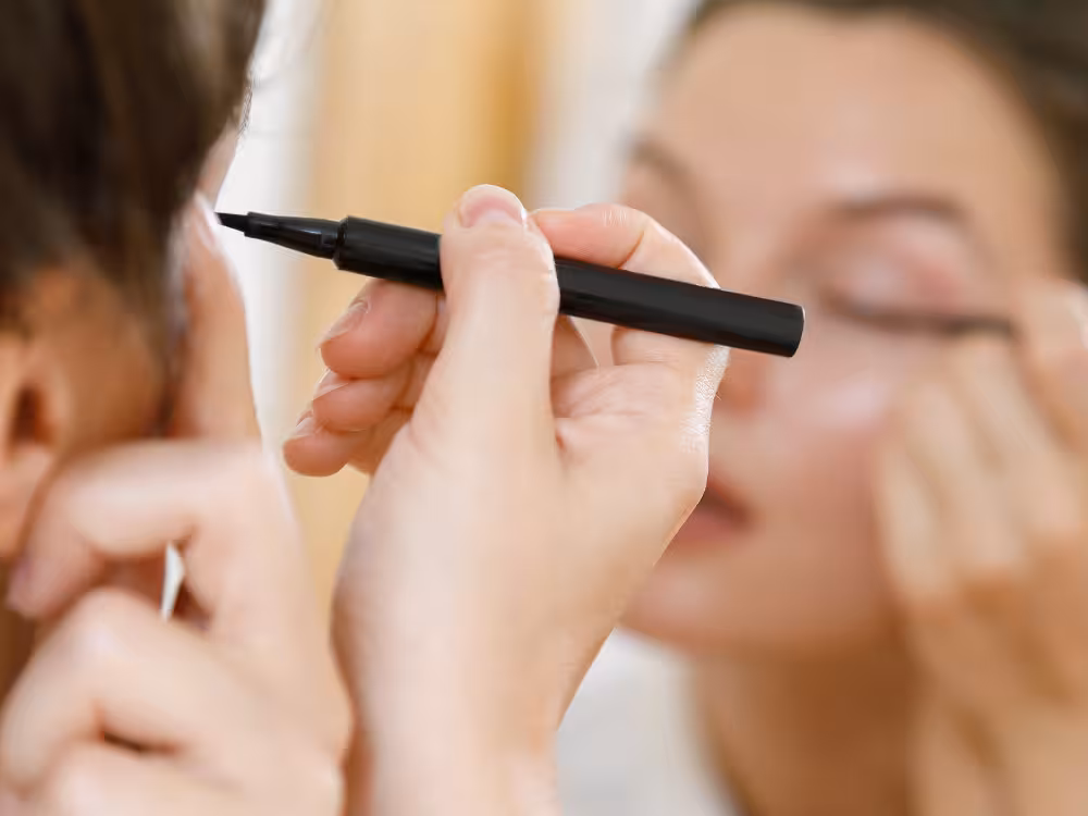 Close-up de mão segurando um delineador líquido preto em caneta, aplicando o produto na pálpebra de uma mulher com olhos fechados, desfocada ao fundo. O foco principal está no produto em destaque no centro da imagem