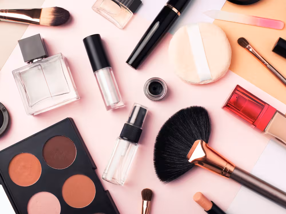 Diversos itens de cosméticos: paleta de contorno, pincéis profissionais, frasco de perfume transparente, rímel e esponja de pó, ilustrando uma rotina de beleza completa.