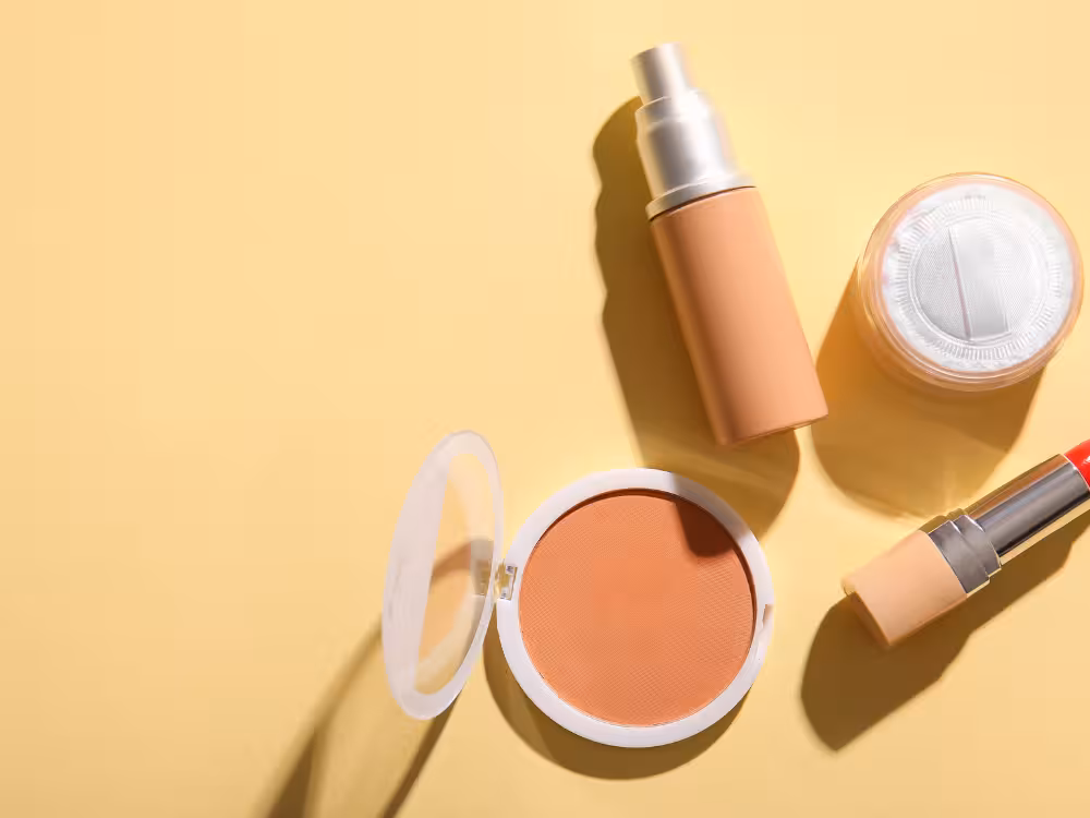 Visão de cima de produtos de maquiagem essenciais, dispostos em uma superfície de cor creme, inclui um blush compacto aberto, um frasco de base, pó translúcido e batom.