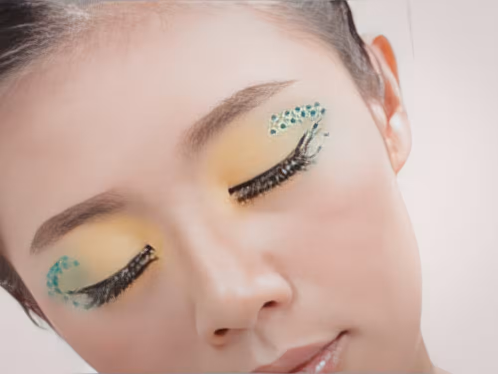 Rosto de mulher jovem asiática com olhos fechados e maquiagem artística: sombra amarela suave nas pálpebras, cílios postiços com textura de penas pretas e strass turquesa agrupados no canto externo de ambos os olhos. Expressão serena, pele clara e fundo neutro claro.