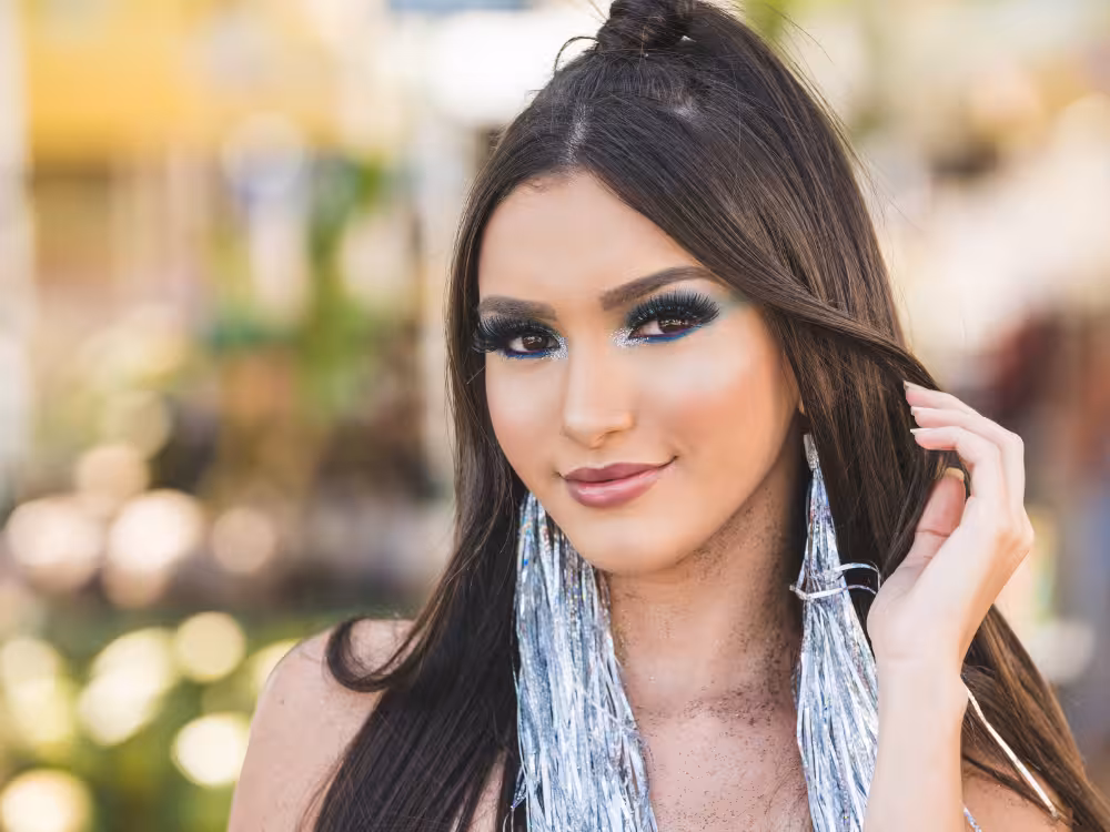 Mulher jovem morena com cabelos lisos escuros semi-presos, olhando levemente para o lado e sorrindo sutilmente. Maquiagem com sombra verde-esmeralda com glitter prateado, cílios postiços volumosos e batom nude rosado. Usa brincos longos de franjas metálicas prateadas. Fundo externo desfocado com tons amarelados e verdes.