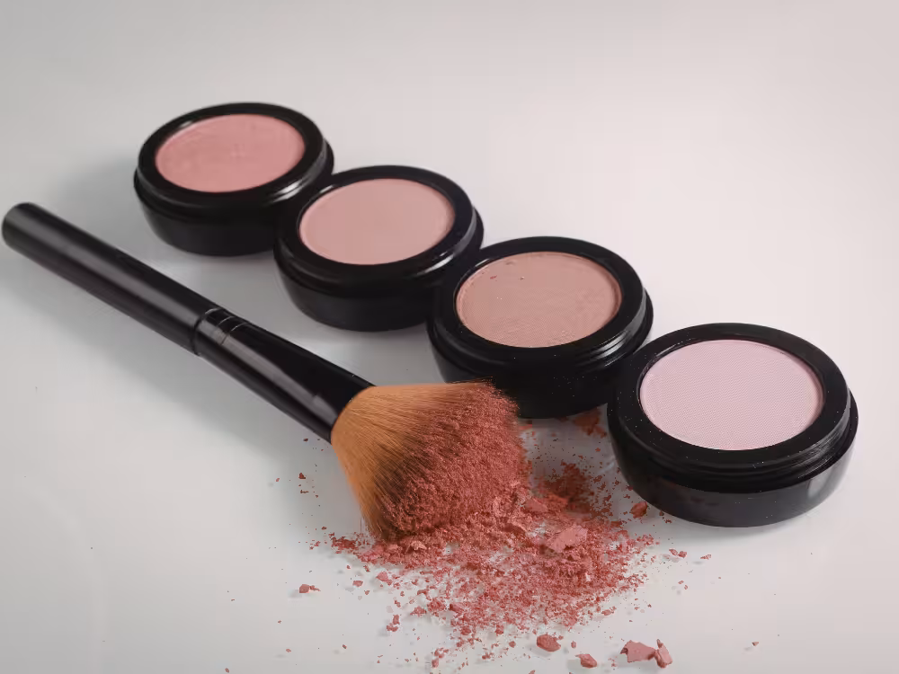 Uma fileira diagonal de quatro potes abertos de blush em pó em tons de rosa e lilás, sobre um fundo branco. Um pincel de maquiagem com cerdas carregadas de pó rosa está deitado sobre a fileira, com um pequeno acúmulo de blush solto na frente