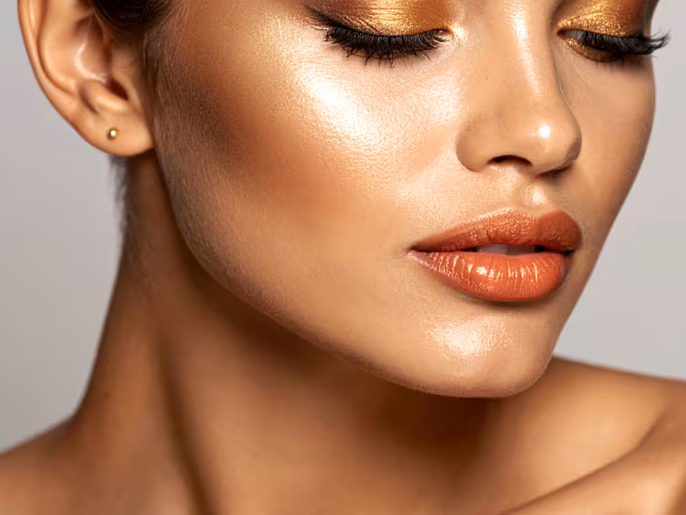 Close-up do rosto de mulher morena com olhos fechados e maquiagem dourada sofisticada: sombra dourada metálica nas pálpebras, cílios postiços volumosos, iluminador intenso nas maçãs do rosto e batom terracota metálico. Brinco pequeno dourado. Pele com efeito glow. Fundo cinza neutro.