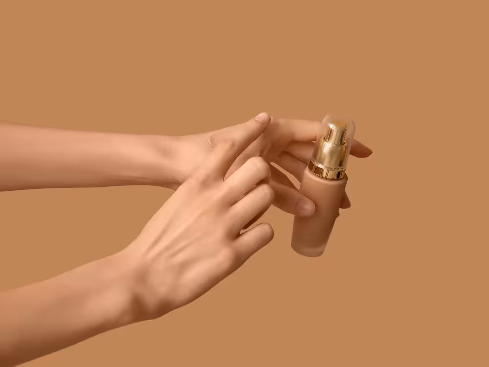 Duas mãos femininas de pele clara segurando um frasco de base líquida bege com bomba dosadora dourada, sem rótulo. Uma mão sustenta o frasco enquanto a outra pressiona o dosador. Fundo terracota bege que harmoniza com o tom do produto.