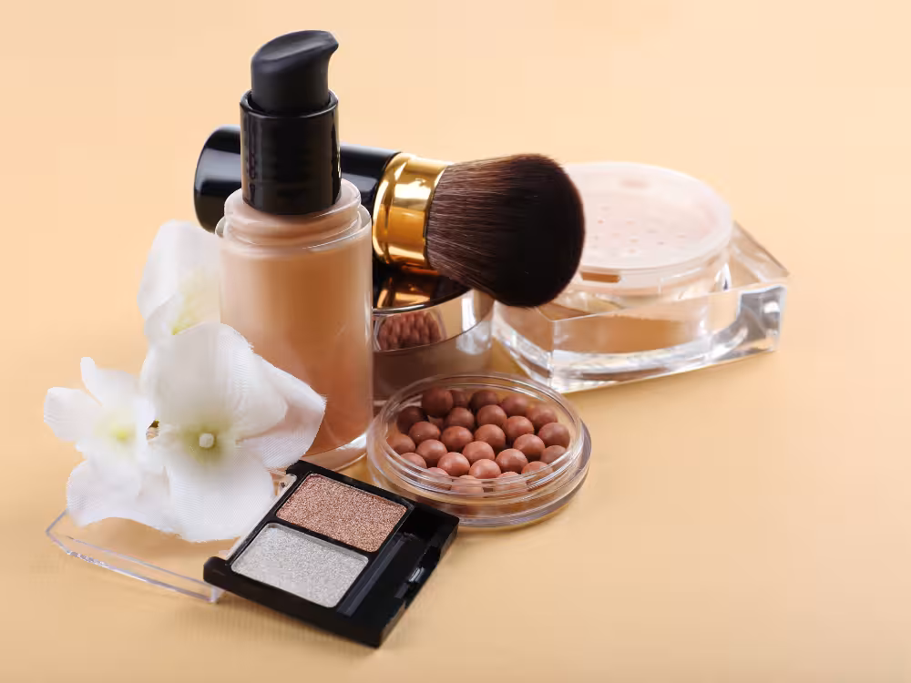 Composição de produtos de maquiagem sobre fundo bege: frasco de base líquida com tampa preta, pincel kabuki com aro dourado, pote transparente de pó solto, estojo redondo com esferas bronzeadoras, paleta duo de sombras em tons nude e dourado, e flor branca decorativa. Estética clean e elegante.