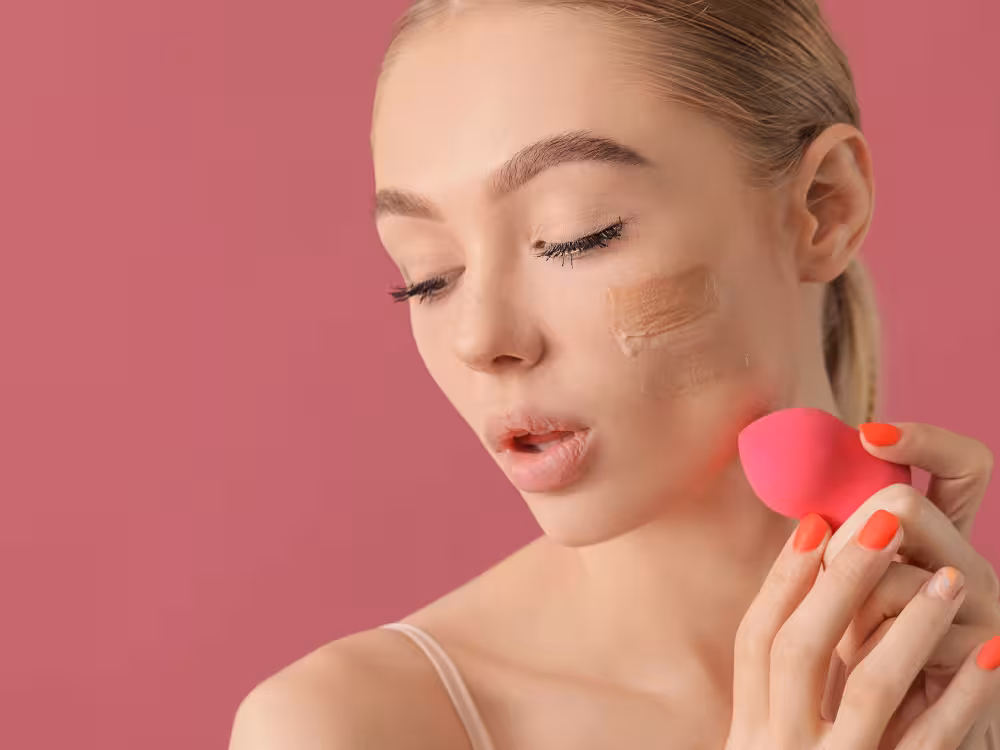 Mulher jovem loira de cabelos presos, olhos semifechados e boca levemente aberta, com listras de base bege aplicadas na bochecha prontas para serem esfumadas. Segura uma esponja de maquiagem rosa coral com unhas vermelhas. Fundo rosa escuro.