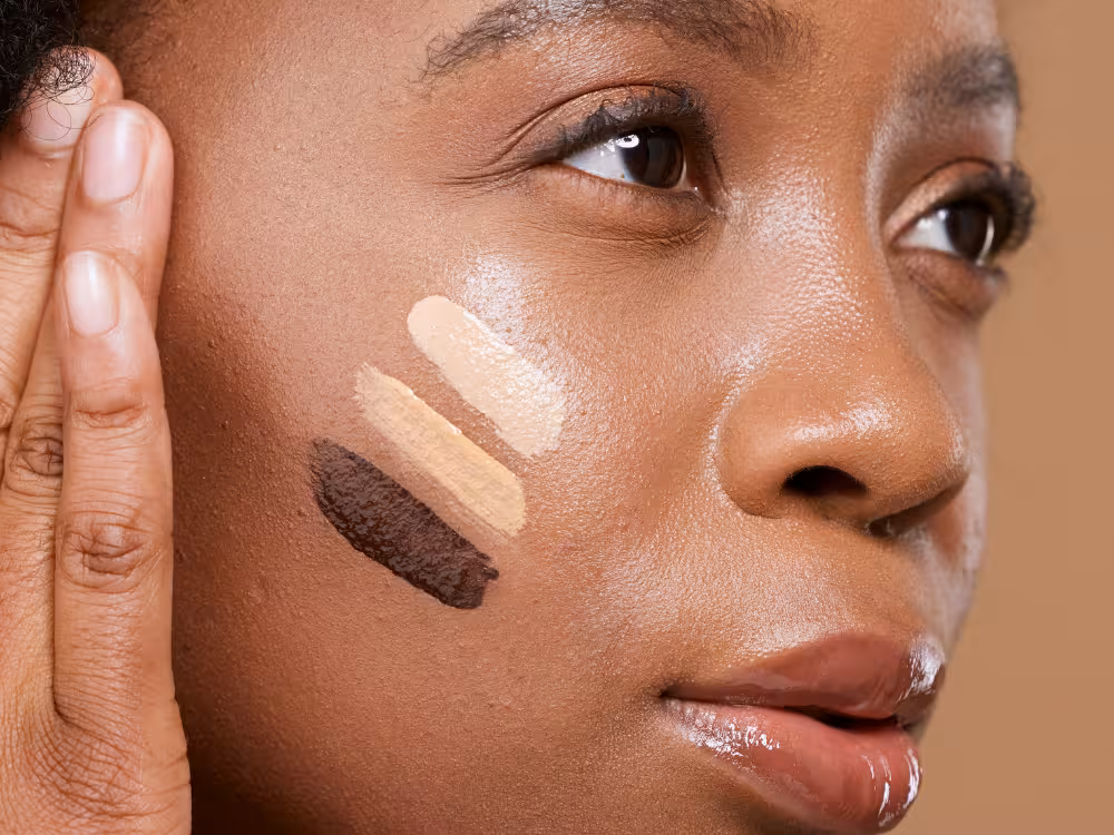 Close-up do rosto de mulher negra com pele escura e luminosa, exibindo três swatches de base em tons diferentes aplicados na bochecha — do mais escuro ao mais claro, em marrom escuro, bege médio e bege claro — para comparação de tonalidades. Mão apoiada na lateral do rosto. Fundo bege neutro.