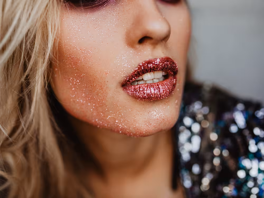 Close-up do rosto de mulher loira com maquiagem festiva: batom vermelho com glitter metálico e glitter prateado fino espalhado pelas bochechas e queixo. Sombra vinho visível no topo da imagem. Roupa com lantejoulas metálicas coloridas. Fundo cinza desfocado.
