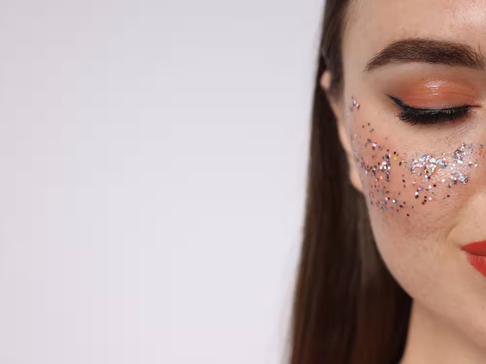 Metade do rosto de mulher jovem de cabelos escuros com olho fechado, sombra coral-rosada, delineador preto, cílios postiços e batom vermelho. Glitter colorido em fragmentos prateados, vermelhos e multicoloridos espalhados pela bochecha. Fundo branco com amplo espaço negativo à esquerda.