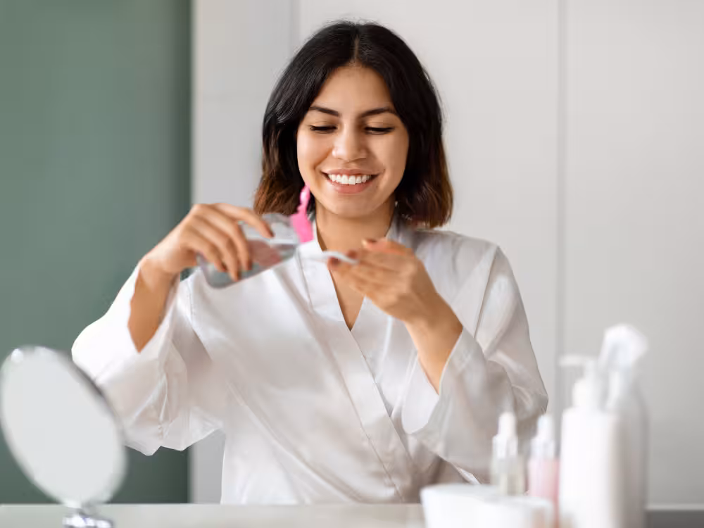 Mulher jovem de cabelos escuros curtos sorrindo enquanto despeja produto de frasco transparente com tampa rosa em um disco de algodão. Veste roupão branco. Ao fundo desfocado, espelho redondo de mesa, frascos de skincare e parede verde-sage. Mesma cena da imagem anterior em ângulo diferente.