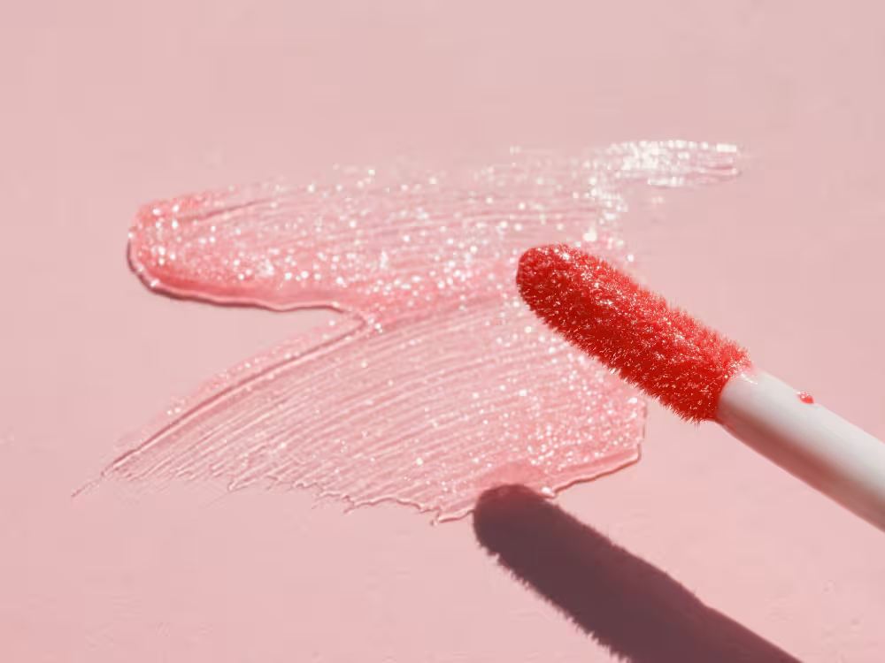 Aplicador de gloss labial com espuma vermelha com glitter sobre mancha esfregada de gloss rosa com brilho espalhada na superfície. Cabo branco e fundo rosa claro. Imagem de textura e swatcher do produto com iluminação natural e sombra projetada.