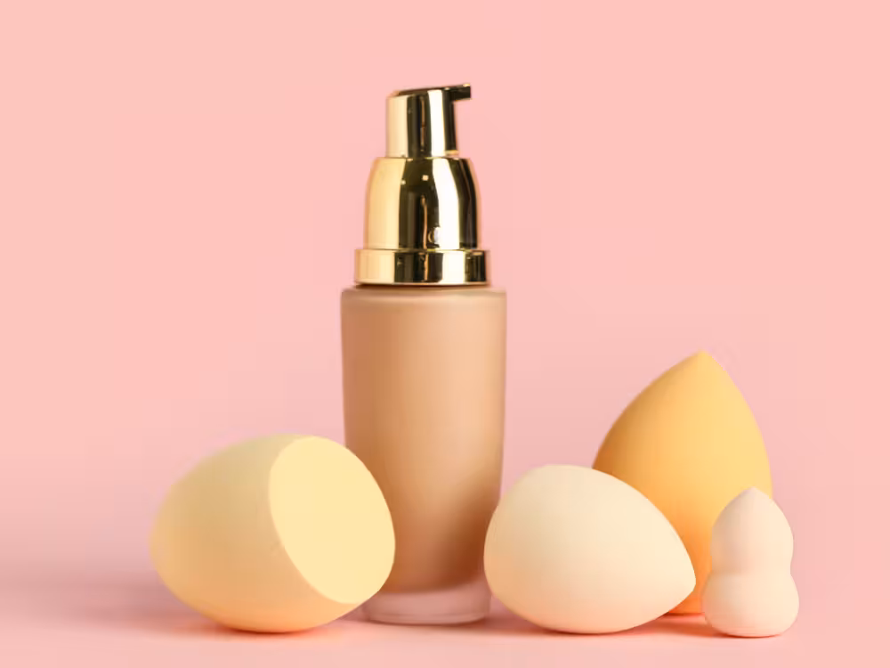 Frasco fosco bege de base líquida com bomba dosadora dourada centralizado, rodeado por quatro esponjas de maquiagem em formatos variados — redonda, oval, gota e formato de boneco de neve — em tons bege e amarelo suave. Fundo rosa claro. Composição de produto com estética minimalista.