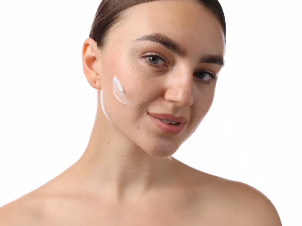 Mulher jovem de cabelos escuros presos, olhos claros e pele natural com algumas manchinhas, sorrindo levemente com creme branco aplicado na bochecha esquerda ainda não espalhado. Ombros descobertos. Fundo branco clean.
