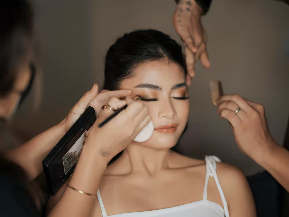 Uma mulher asiática jovem com os olhos fechados recebe maquiagem profissional. Uma maquiadora aplica sombra nos olhos com um pincel, enquanto outra mão segura uma esponja de maquiagem perto de seu rosto. Uma terceira mão com tatuagem aparece acima da cabeça dela. A mulher usa top branco de alcinha e tem cabelo escuro preso