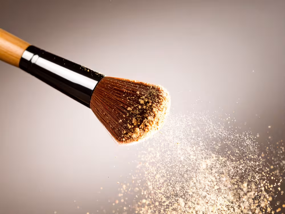 Pincel de maquiagem com cabo de bambu e cerdas marrom-avermelhadas carregadas de pó bege dourado, liberando uma nuvem de partículas iluminadas em movimento para o lado direito. Fundo cinza com iluminação dramática realçando o efeito de dispersão do produto.