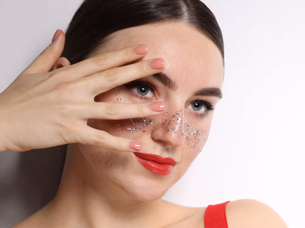 Mulher jovem de cabelos escuros presos e olhos azuis, cobrindo parte do rosto com a mão de unhas rosadas. Glitter prateado e multicolorido espalhado pelo nariz e bochechas simulando sardas. Batom vermelho e blusa vermelha. Fundo branco neutro.