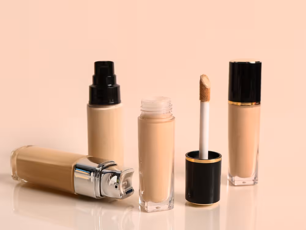 Quatro produtos de base e corretivo em tons bege dispostos sobre superfície reflexiva: frasco deitado com tampa prata, frasco com bomba preta, corretivo líquido aberto com aplicador de esponja bege, e frasco quadrado com tampa preta e aro dourado. Fundo rosé claro. Composição de produtos sem rótulo.