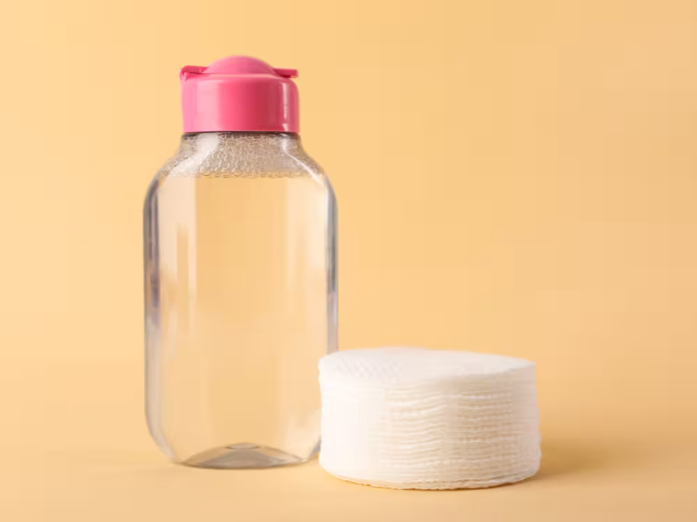 Frasco transparente quadrado com tampa flip-top rosa contendo líquido levemente amarelado, ao lado de uma pilha de discos de algodão brancos empilhados. Fundo amarelo bege claro. Composição simples de produtos de skincare para destaque dos itens.