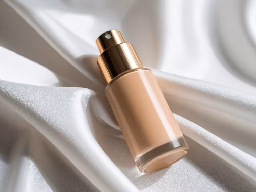 Frasco de base líquida bege com bomba dosadora dourada, sem rótulo, apoiado na diagonal sobre tecido de cetim branco com dobras suaves. Iluminação natural com sombras delicadas. Estética luxuosa e minimalista.
