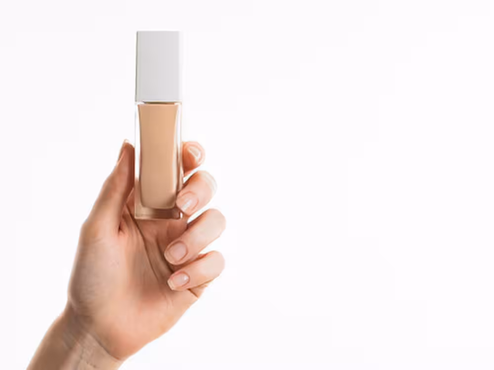 Mão feminina de pele clara segurando um frasco transparente de base líquida bege com tampa branca quadrada, sem rótulo visível. Fundo branco clean. Imagem com estética minimalista para destaque do produto.