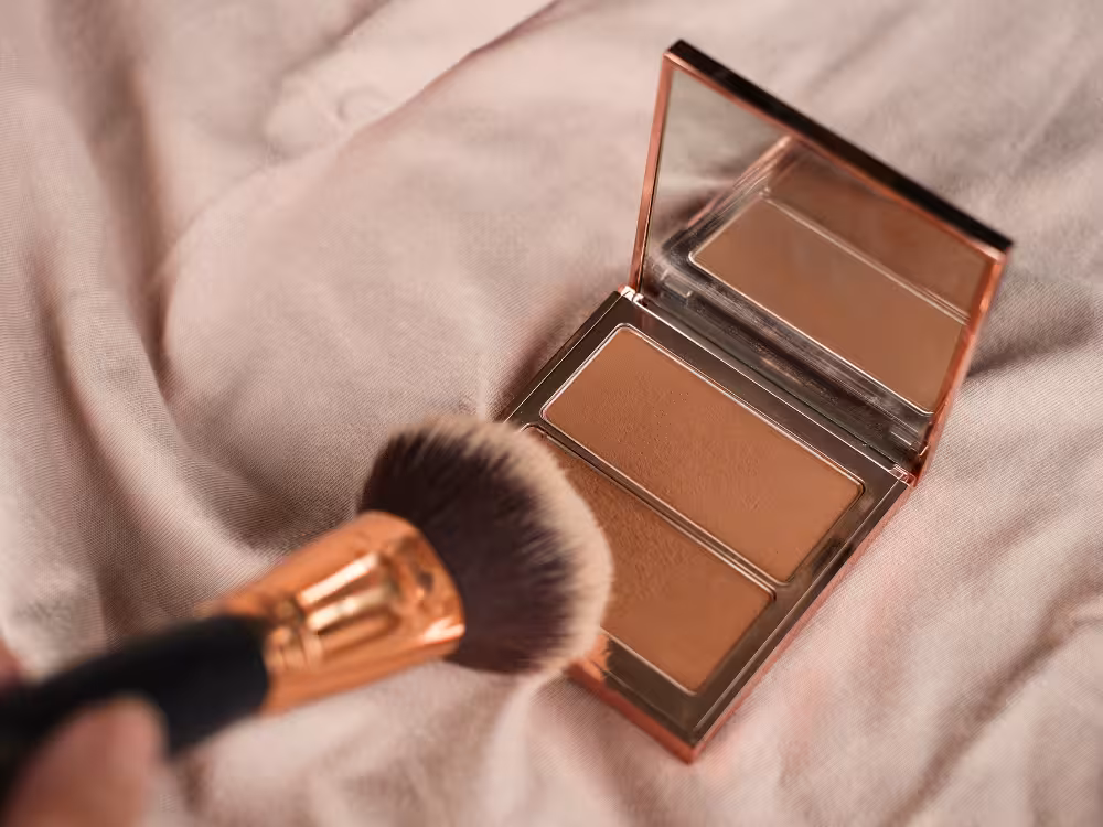 Um pincel de maquiagem com cabo rose gold é segurado próximo a uma paleta de contorno aberta. A paleta metálica rose gold contém dois tons de pó marrom em um estojo com espelho. O conjunto está apoiado sobre um tecido macio e claro em tons pastel.