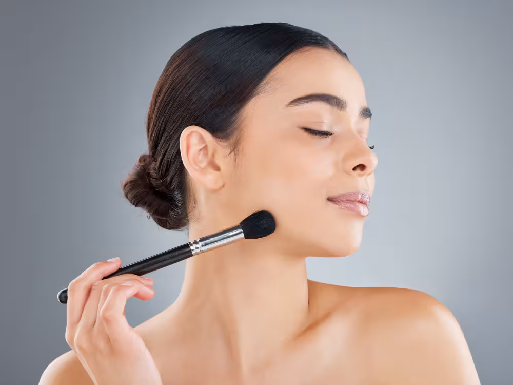 Uma mulher de pele morena clara e cabelo escuro preso em um coque aparece de perfil, com os olhos fechados e um leve sorriso. Ela segura um pincel de maquiagem preto com cabo prateado contra a bochecha, como se estivesse aplicando blush ou contorno. Fundo cinza neutro.