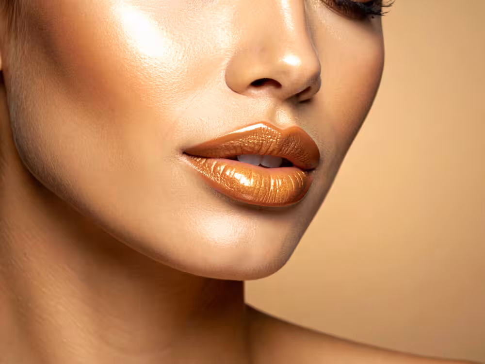 Close-up do rosto de mulher com pele morena dourada e iluminada, exibindo batom marrom-caramelo metálico com acabamento glossy. Maquiagem com efeito glow intenso nas maçãs do rosto e nariz. Fundo bege neutro.