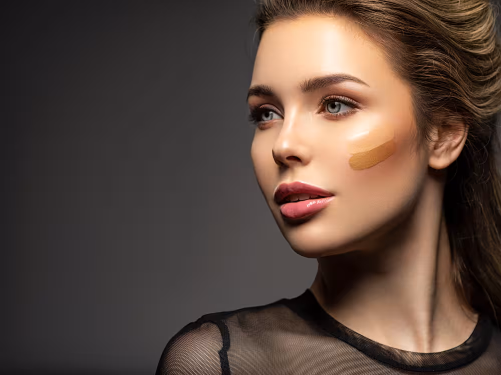 Mulher jovem de cabelos castanhos soltos e olhos azuis com expressão sofisticada, swatch de base bege dourado esfumado na bochecha, batom vinho escuro e maquiagem editorial com iluminador. Blusa de tule preta. Fundo cinza escuro com amplo espaço negativo à esquerda. Iluminação dramática de estúdio.