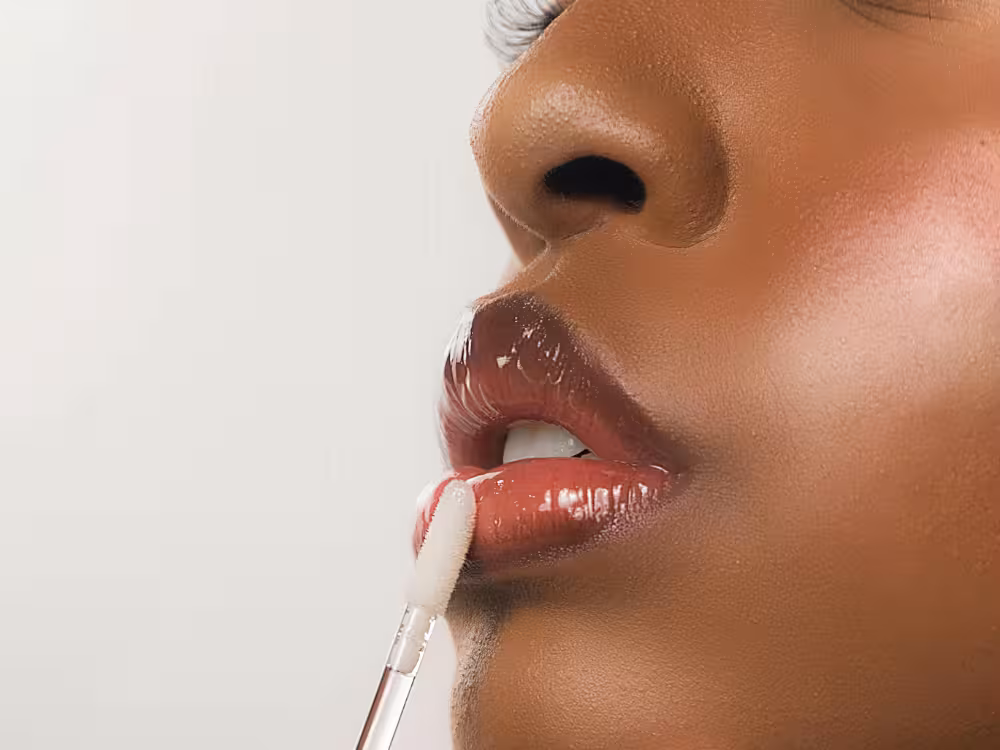 Close-up lateral do rosto de mulher negra com pele escura e luminosa, aplicando gloss transparente sobre batom vermelho escuro com acabamento úmido. O aplicador de espuma aparece no canto inferior da imagem. Fundo cinza claro neutro.
