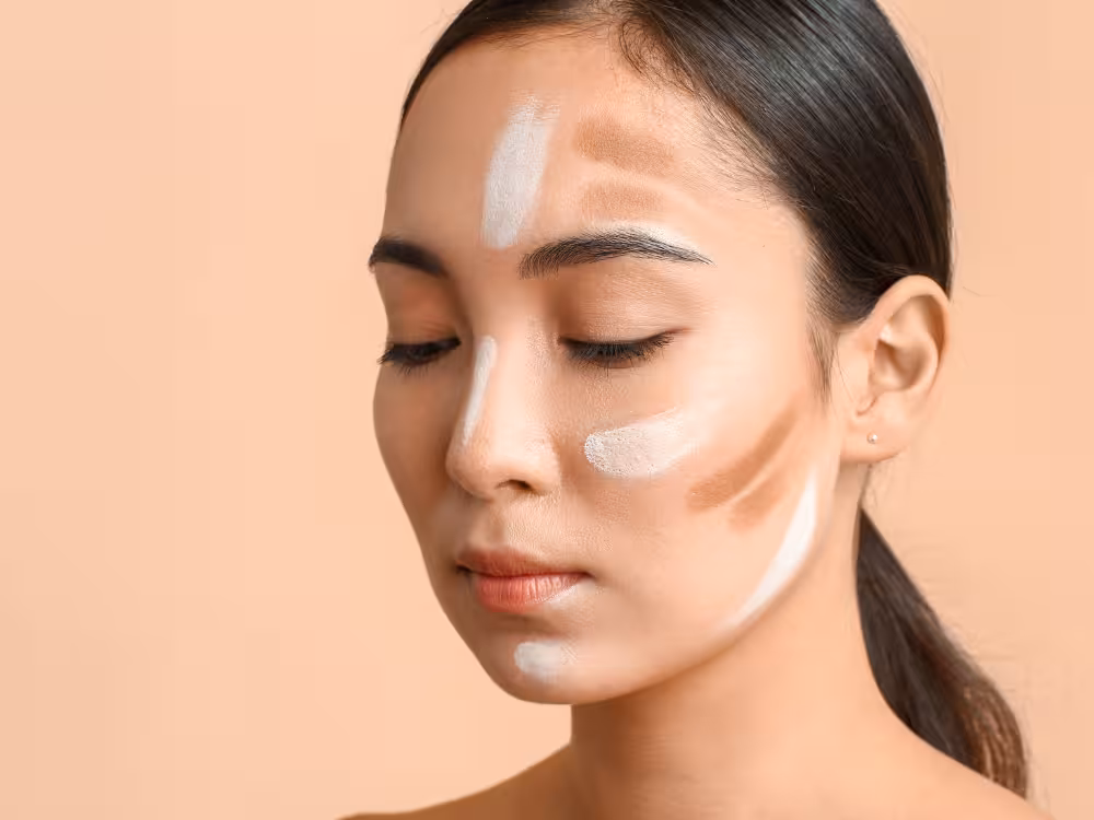 Uma mulher asiática de pele clara e cabelo escuro preso em rabo de cavalo aparece em close, com os olhos fechados. Seu rosto tem produtos de maquiagem aplicados em listras para contouring e highlighting: tons claros (branco/bege) na testa, nariz, maçãs do rosto e queixo, e tons mais escuros (marrom) nas laterais da testa e bochechas. Fundo em tom bege suave.