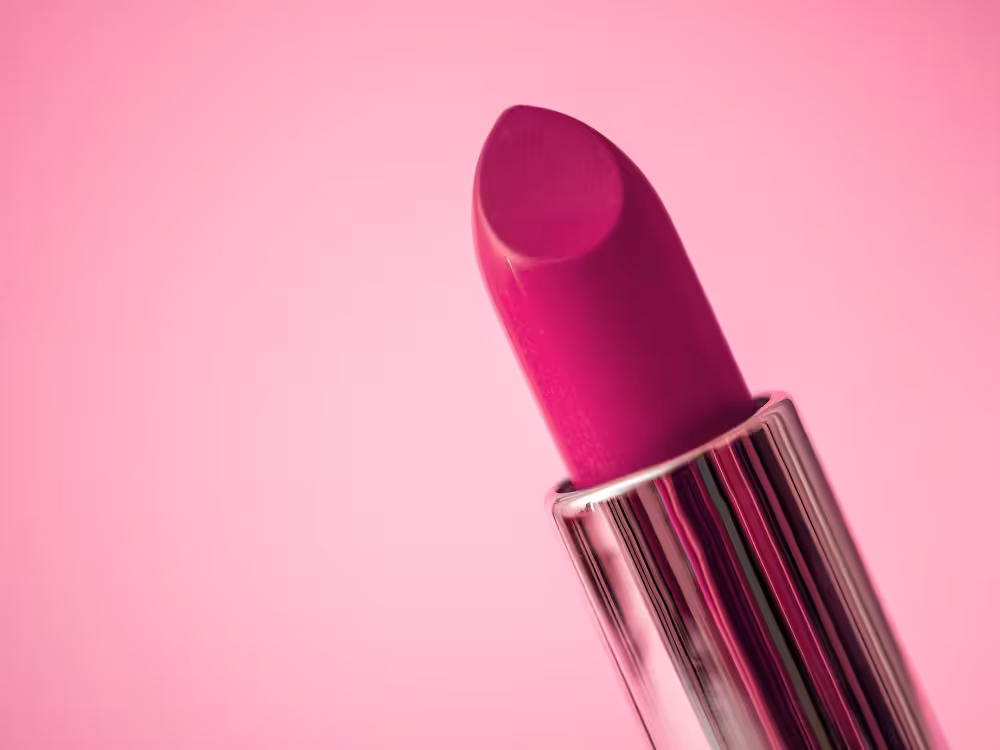 Close-up de batom matte em tom magenta vibrante com embalagem metálica prateada canelada, sobre fundo rosa claro.