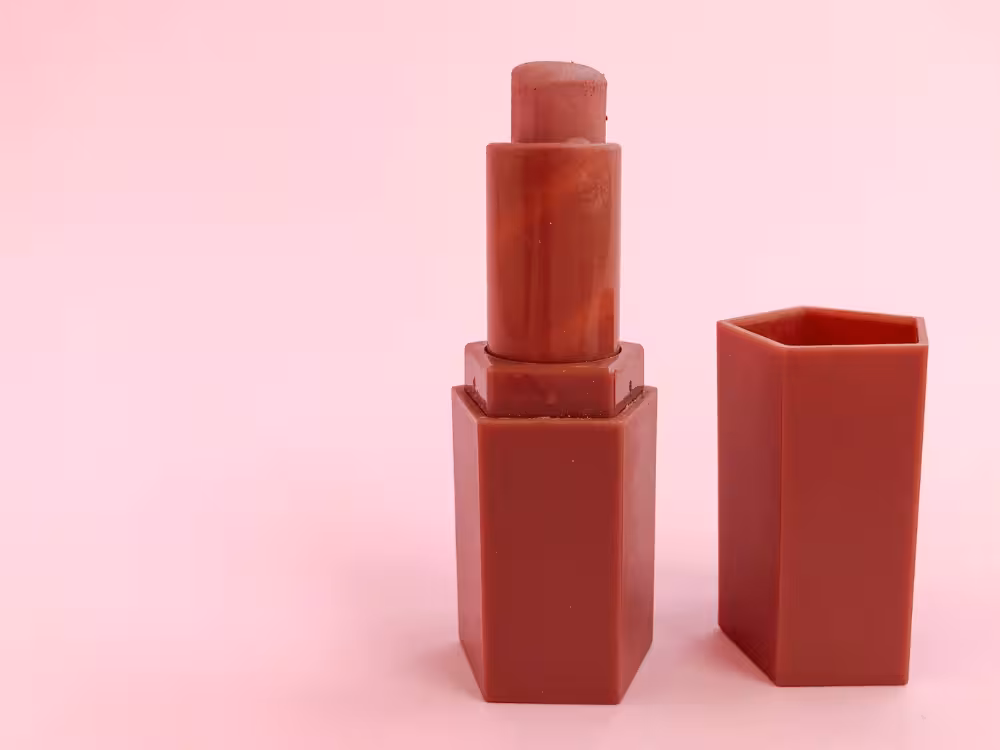 Batom aberto em tom vinho escuro com embalagem hexagonal fosca na mesma cor, ao lado da tampa, sobre fundo rosa claro. Design minimalista e monocromático com formato de base sextavada.