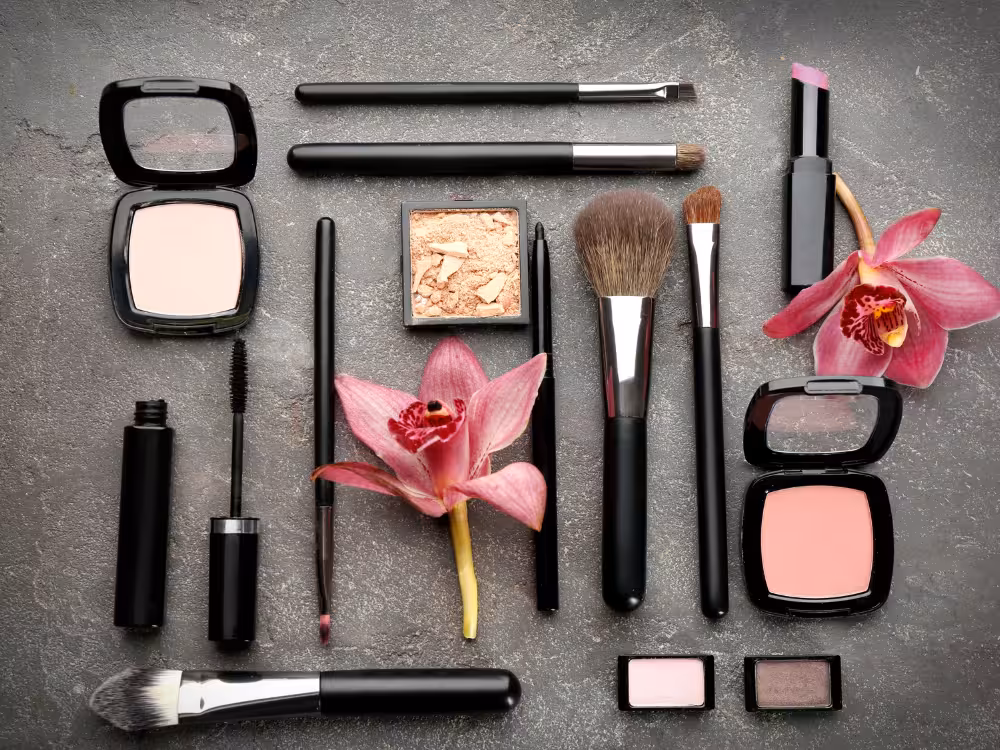 Flat lay de produtos e acessórios de maquiagem com embalagens pretas sobre superfície de concreto cinza escuro: pó compacto, rímel, lápis labial, pincel de sobrancelha, sombra dourada, três pincéis faciais, batom, blush rosa, duas sombras individuais em rosa e marrom, e duas orquídeas rosas como elemento decorativo.