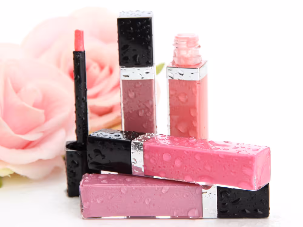 Conjunto de produtos labiais com gotas d'água sobre as embalagens pretas com detalhes prateados: dois glosses líquidos em tons rosa e malva, dois batons em rosa pink e lilás, e um lápis labial rosa. Rosa desfocada ao fundo. Fundo branco.