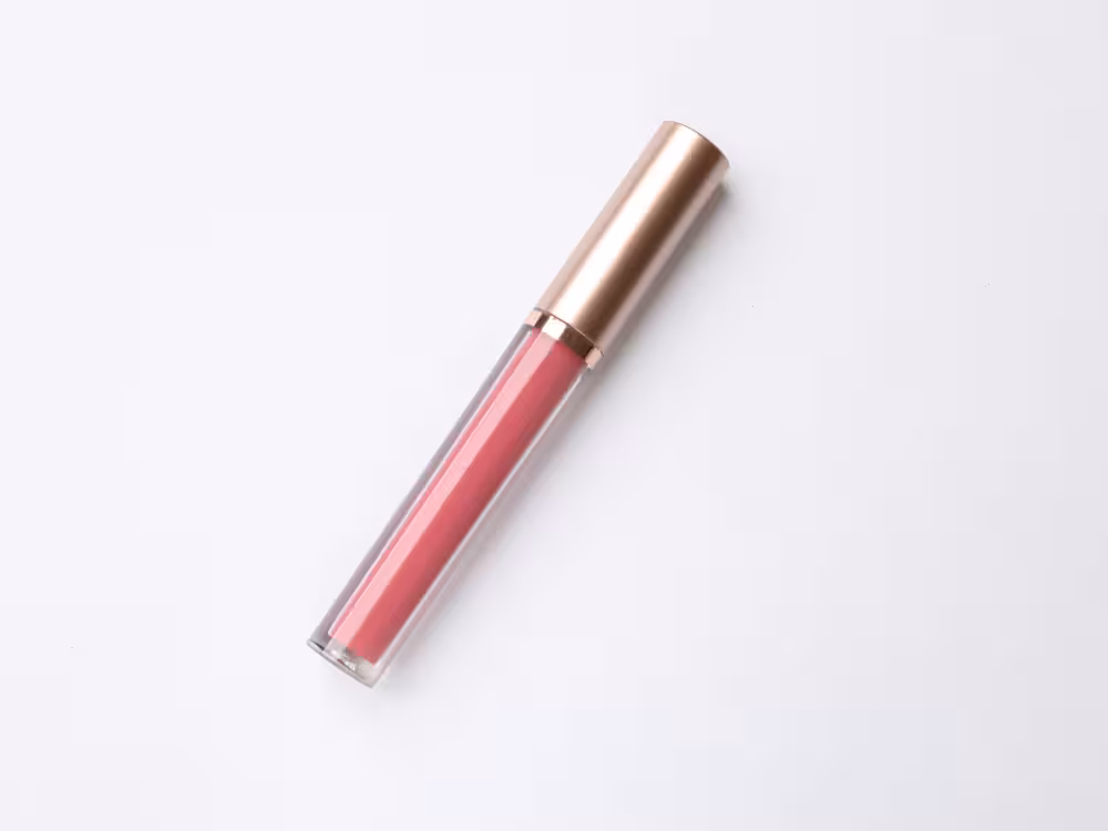 Gloss labial em tom rosa médio com frasco transparente retangular e tampa dourada rose gold, deitado na diagonal sobre fundo branco. Sem rótulo, composição minimalista com estética luxuosa.