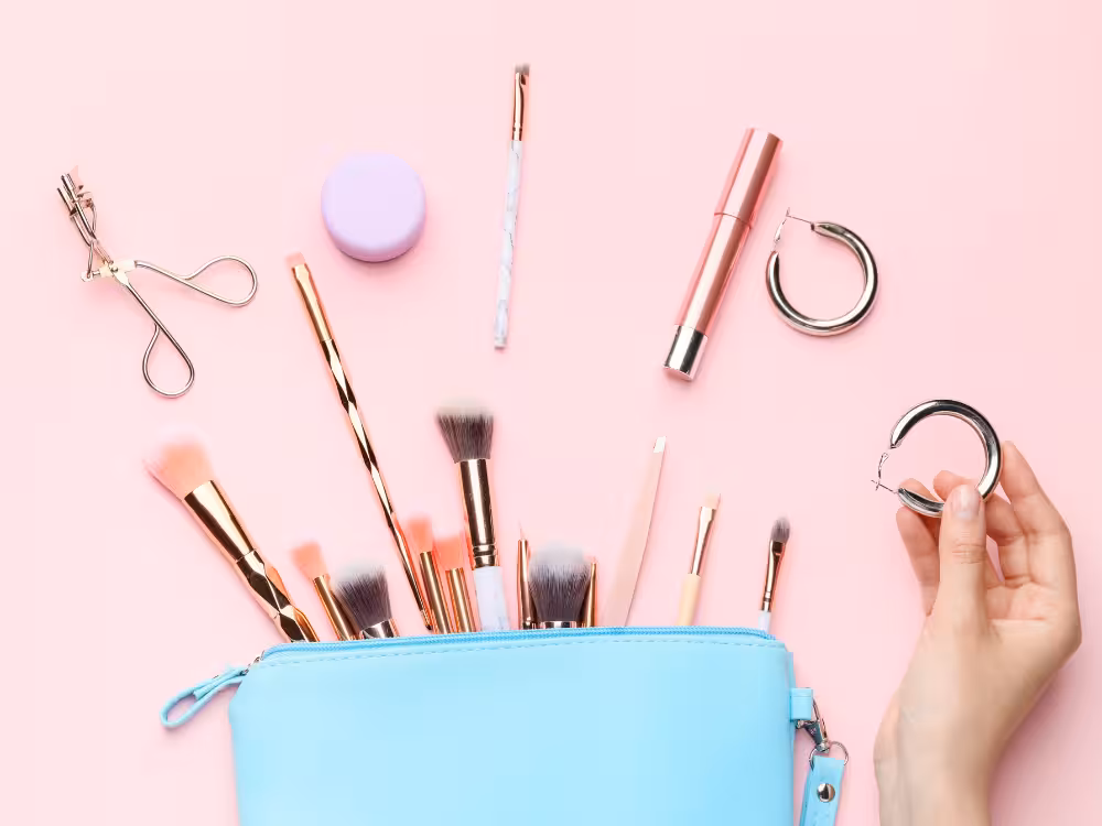 Nécessaire azul claro aberta com pincéis de maquiagem rose gold e cabo marmorizado espalhados sobre fundo rosa. Ao redor: curvex metálico, pote lilás, batom rosa, pinça e mão segurando um par de argolas prateadas.