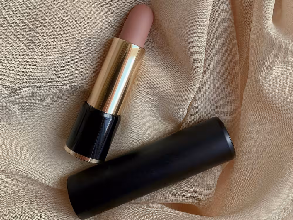 Batom aberto em tom nude rosado com acabamento matte, embalagem preta com aro dourado, apoiado ao lado da tampa sobre tecido de cetim bege com dobras suaves. Composição elegante com estética luxuosa.