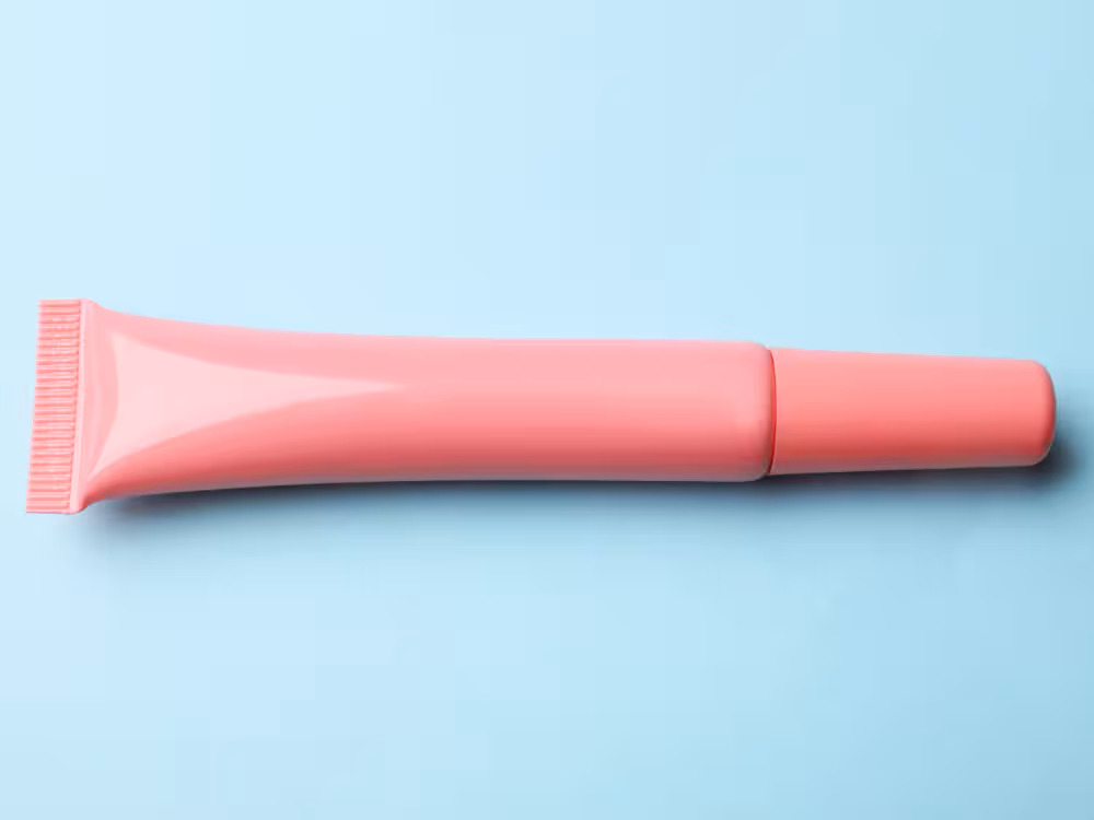 Bisnagas de gloss labial rosa salmão com aplicador de pente na ponta, deitada sobre fundo azul claro. Embalagem monocromática sem rótulo em composição minimalista.