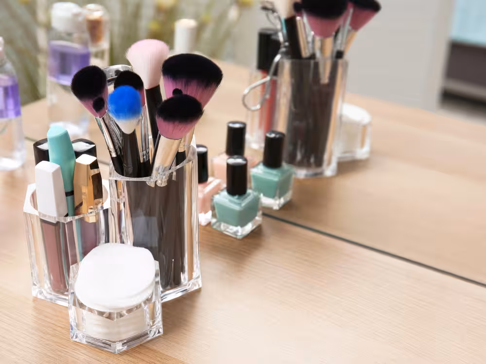 Organizador acrílico transparente sobre penteadeira de madeira clara com pincéis coloridos, batons, gloss e esponja de maquiagem. Ao lado, esmaltes em verde e rosa enfileirados. Espelho ao fundo reflete os itens. Ambiente claro e organizado.