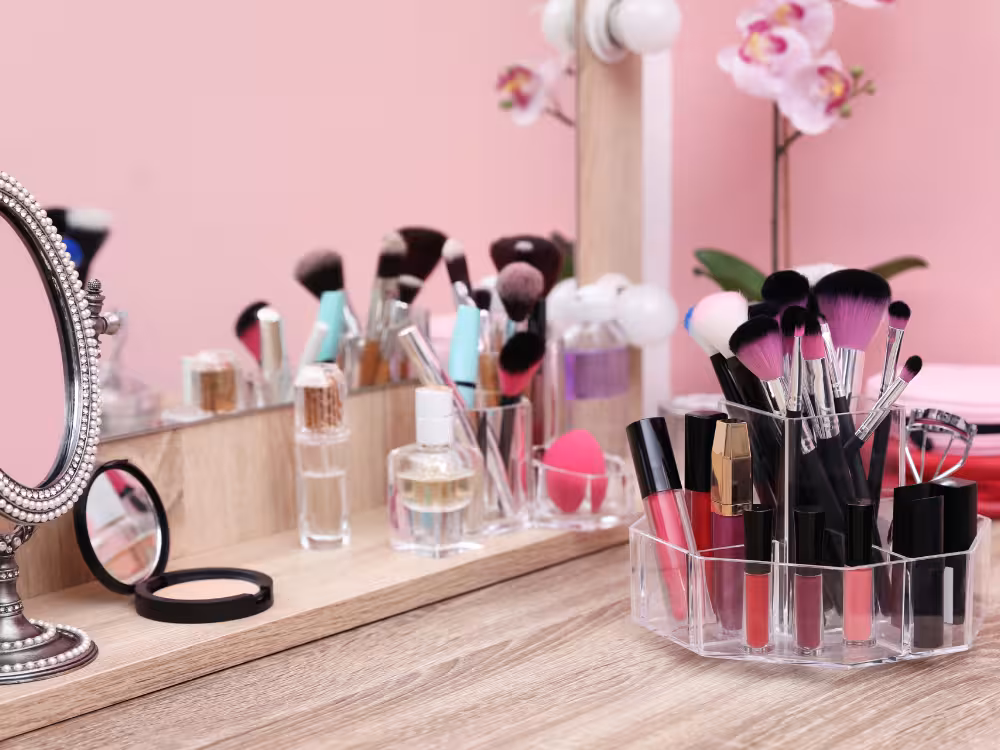 Penteadeira de madeira clara com organizador acrílico giratório contendo pincéis, glosses e batons rosa, bandeja de madeira com pincéis, frascos de perfume e esponja pink ao fundo, espelho redondo com moldura prateada e pó compacto à esquerda. Orquídeas rosas e parede rosa ao fundo.