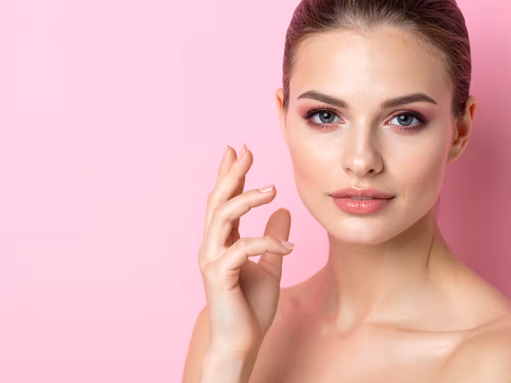 Mulher branca de cabelos castanhos com maquiagem rosa nos olhos. Ambiente com fundo rosa.