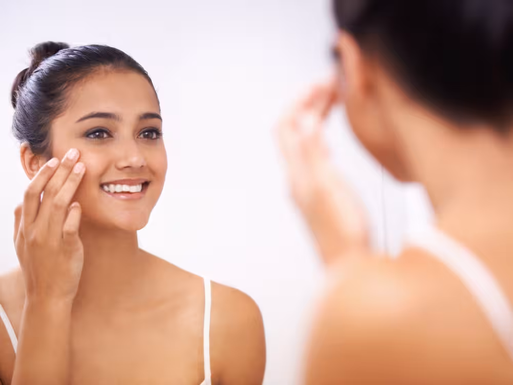 Mulher branca com cabelo preto em ambiente de fundo branco fazendo uma limpeza de pele com um sorriso no rosto