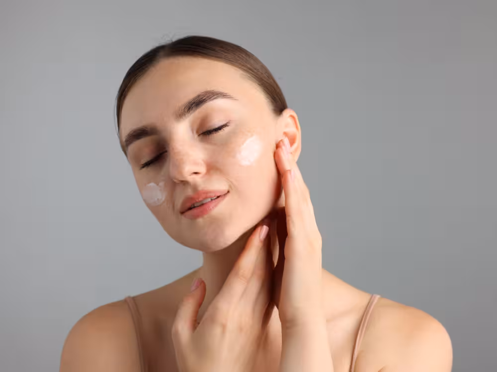 Mulher de cabelos castanhos, com as duas mãos no rosto, com creme hidratante nas maçãs do rosto, de olhos fechados num ambiente com fundo cinza.