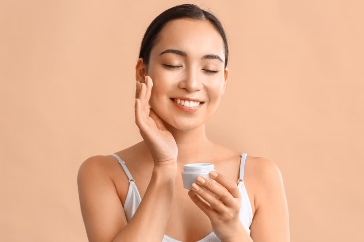 Mulher branca com cabelo escuro passando hidratante no rosto, num ambiente com fundo branco.
