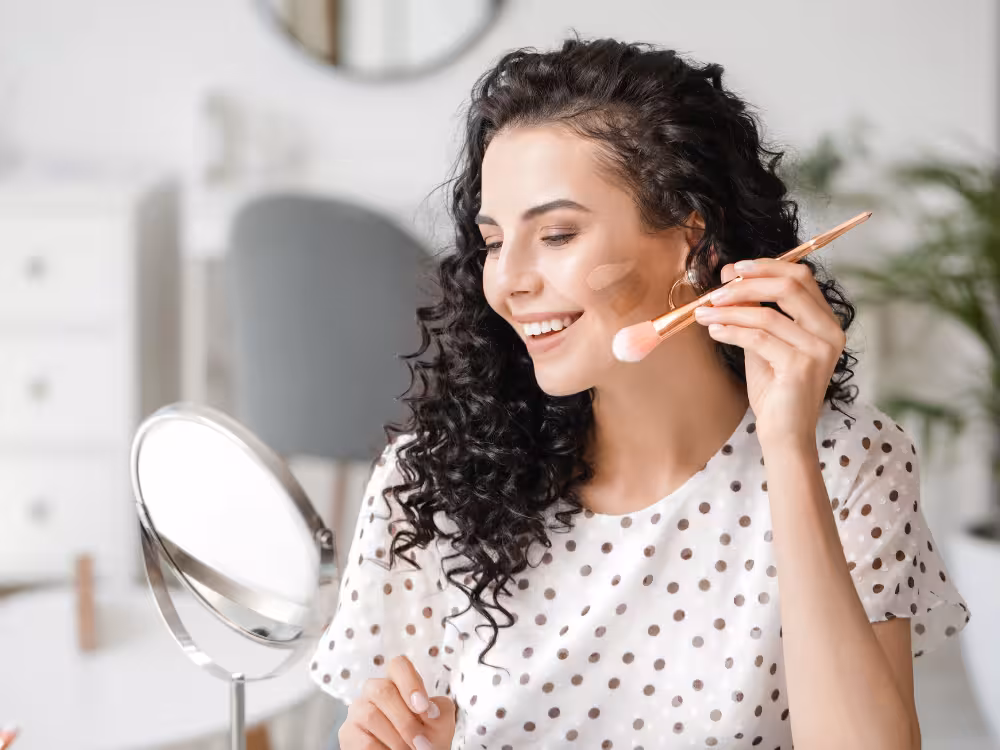 Mulher com cabelo encaracolado e preto, com roupa branca com bolinhas pretas passando base no rosto.
