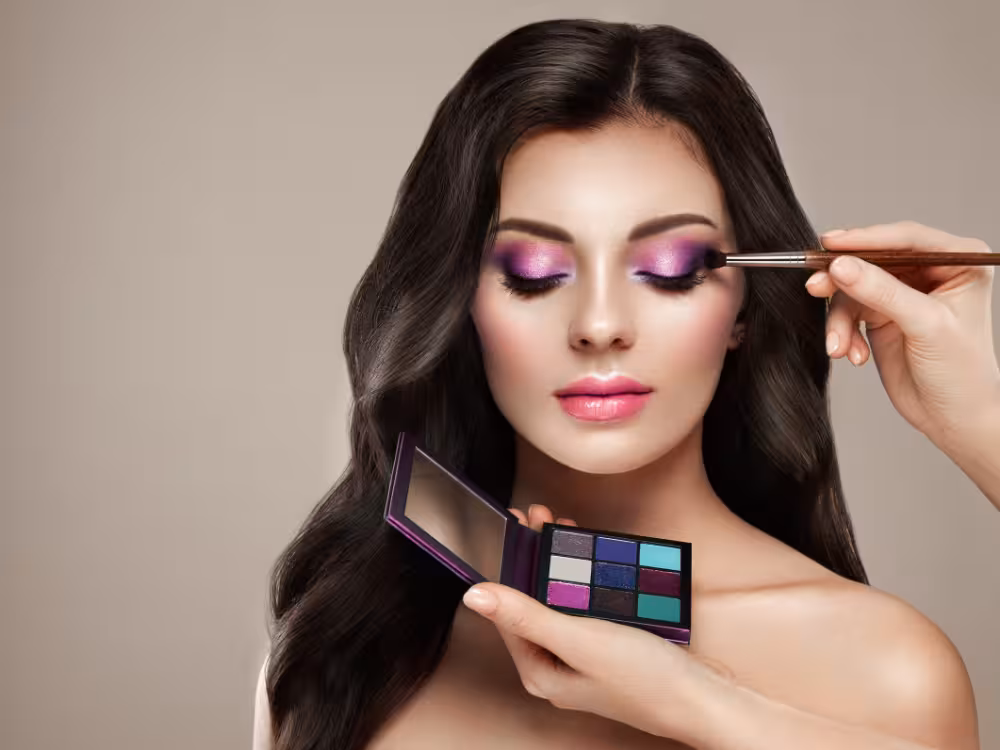Mulher branca do cabelo castanho, lábios rosa fazendo maquiagem rosa nos olhos com uma maquiadora que segura um pincél e uma paleta de maquiagem. Ambiente com fundo bege.